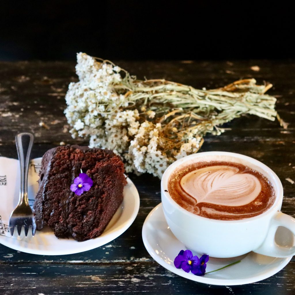 a cup of barista coffee and a chocolate cake on a rustic table with flowers