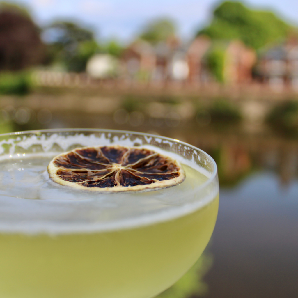 A green cocktail decorated with dehydrated orange against a backdrop of a river with houses in the distance